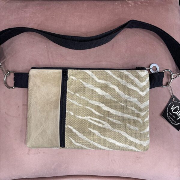 Custom tan and cream animal print fabric crossbody bag with tan leather and black vertical front zipper. Silver circle and hook hardware attaches strap-silver charm on zipper-black kcity hangtag- pink background.