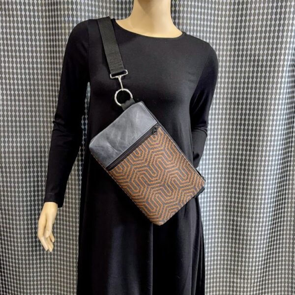 Mannequin wearing black dress with custom rust and charcoal print crossbody bag with front vertical zipper and black leather strip. Silver circle and hook hardware attached at strap. Backdrop grey print curtain.