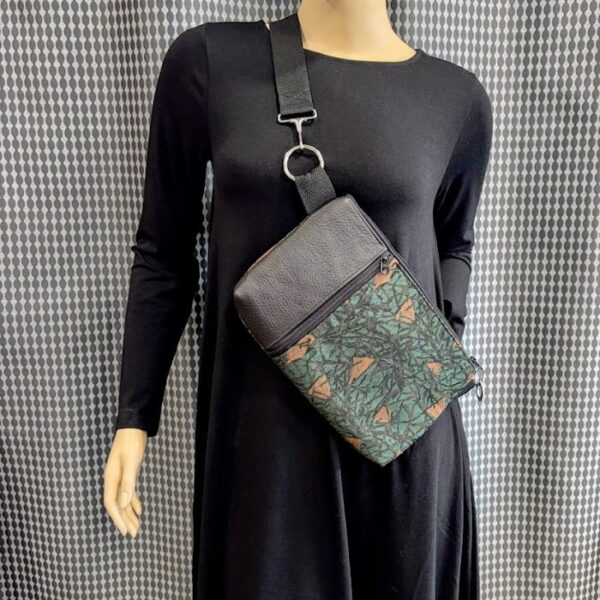 Mannequin wearing black dress displaying custom crossbody bag with forest green fabric printed with brown triangles and black scribble lines with front vertical zipper and black leather strip-silver circle and hook hardware attached at strap. Grey print curtain backdrop.