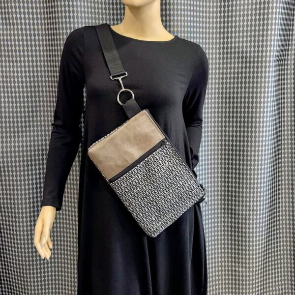 Mannequin wearing black dress-custom crossbody bag with black, tan, white woven design-black vertical zipper-tan leather strip. Silver circle and hook hardware. Grey print curtain background.